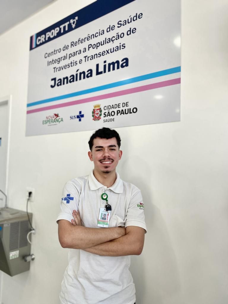 Na imagem, Gustavo está com os braços cruzados em frente a uma placa do CR POP TT. Ele é um jovem de 20 anos, branco, cabelos escuros, de bigode e cavanhaque. Ele usa uma camiseta branca com símbolos do SUS e um crachá com sua foto e nome. Na sua lateral esquerda, aparece parte de um bebedouro preso à parede